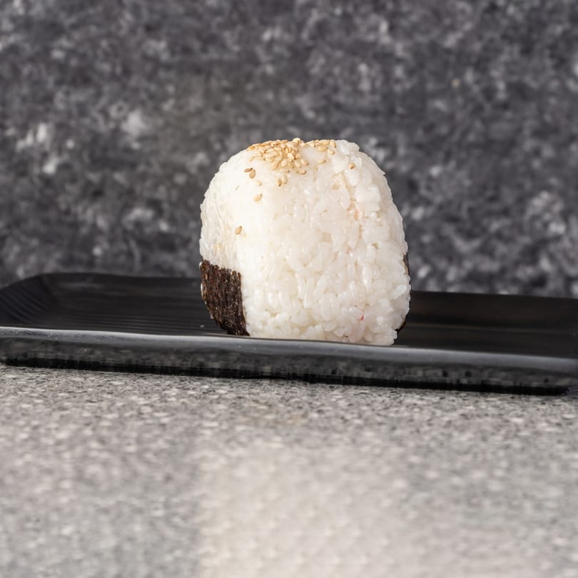 Onigiri rice ball with nori and sesame seeds on a black plate, against a grey background.