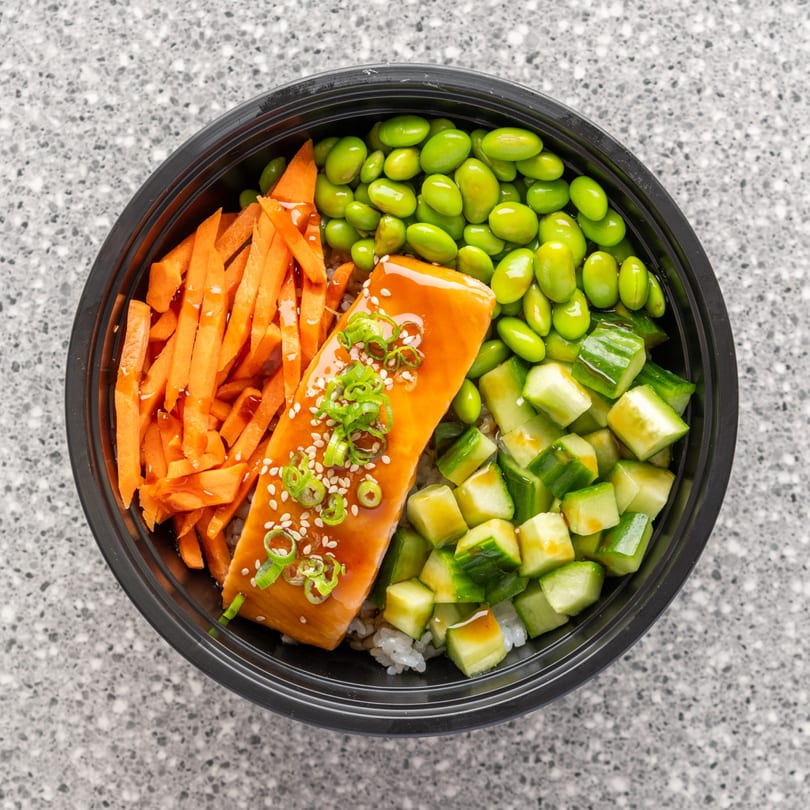 A salmon poke bowl with edamame, carrots, and cucumbers in a black bowl.